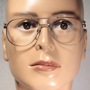 May include: Aviator-style spectacles with a gold-coloured metal frame. The spectacles have a double bridge and clear lenses. The image shows the spectacles on a mannequin head with brown eyes and a neutral skin tone. A classic style.