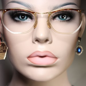 May include: A close-up of a mannequin wearing gold-rimmed glasses with a tortoiseshell brow bar. The glasses have clear lenses and are on a mannequin with blue eyes and pink lips. A gold tag with the text "ETOILE ELITE" hangs from the glasses.