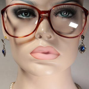 Può includere: Una testa di manichino con grandi occhiali con montatura marrone. Gli occhiali hanno una forma classica e oversize. Il manichino indossa anche orecchini con pendenti di pietre preziose blu. Il manichino ha labbra carnose e lunghe ciglia.