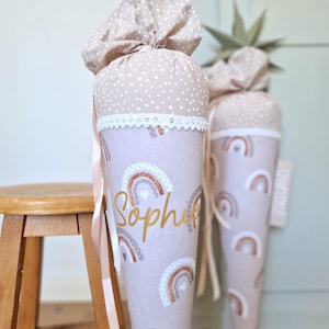 Könnte beinhalten: Zwei kegelförmige Geschenktüten aus Stoff mit beigem Hintergrund und weißen Punkten im oberen Bereich sowie einem Regenbogenmuster in Braun-, Weiß- und glitzernden Kupfertönen. Der Name Sophie ist in goldener Schreibschrift aufgedruckt.