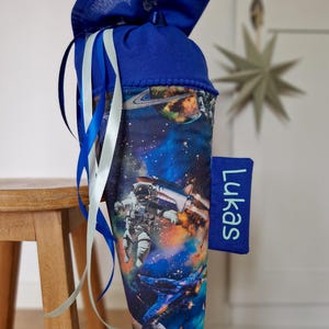 Op de afbeelding: Een blauwe en zwarte stoffen kegel met een ruimtethema print. De kegel heeft een blauw lint en een wit lint eromheen gebonden. De kegel heeft een label met de tekst "Lukas".