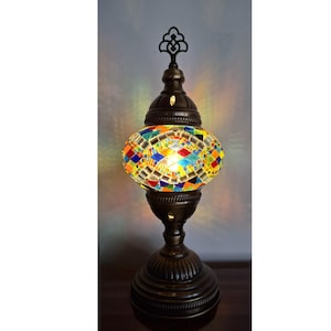May include: A decorative table lamp with a mosaic glass shade. The lamp features a bronze-colored base and a colorful, multi-faceted glass shade. The shade is composed of various colors, including red, blue, yellow, and green. The lamp has an ornate, decorative top.