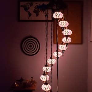 May include: A tall, spiral-shaped floor lamp with a metal base and a series of colorful, mosaic-style glass globes. The globes are lit from within, casting a warm glow. The lamp is in a room with a world map and a dartboard on the wall.