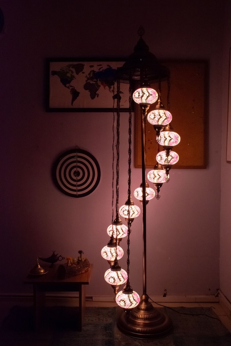 May include: A tall, spiral-shaped floor lamp with a metal base and a series of colorful, mosaic-style glass globes. The globes are lit from within, casting a warm glow. The lamp is in a room with a world map and a dartboard on the wall.