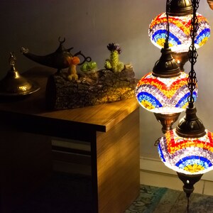 May include: A three-tiered floor lamp with colorful mosaic glass shades. The lamp is lit and casts a warm glow. The lamp is on a wooden table with a decorative brass lid and a genie lamp.