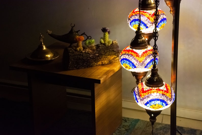 May include: A three-tiered floor lamp with colorful mosaic glass shades. The lamp is lit and casts a warm glow. The lamp is on a wooden table with a decorative brass lid and a genie lamp.