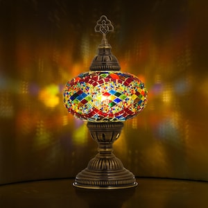 May include: A decorative table lamp with a mosaic glass shade in a variety of colors, including red, blue, green, and yellow. The lamp has a bronze-colored metal base and a decorative finial at the top. The lamp is lit, casting colorful light.