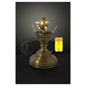 May include: A gold-toned metal lamp with a round, ornate base and a decorative top with a cluster of small, white LED lights. The lamp is powered by two AA batteries, which are shown in the image.