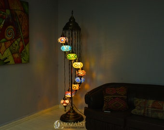 May include: A tall, spiral-shaped floor lamp with a gold metal base and a series of colorful, round, mosaic glass globes. The globes are lit from within, casting a warm glow. The lamp is in a room with a brown leather couch and a painting on the wall.