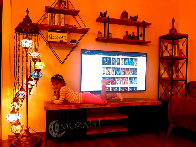 Może przedstawiać: Mała dziewczynka leży na drewnianym stole przed telewizorem, na kt&oacute;rym wyświetlana jest strona internetowa z kolorowymi lampami mozaikowymi. Pok&oacute;j jest ozdobiony drewnianymi p&oacute;łkami i dużą, kolorową lampą podłogową z mozaiki.