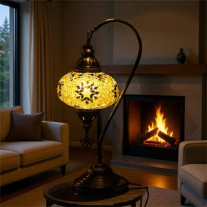 May include: A decorative floor lamp with a mosaic glass shade in shades of yellow and gold. The lamp has a dark bronze metal base and a curved arm. The lamp is near a fireplace with a burning fire.