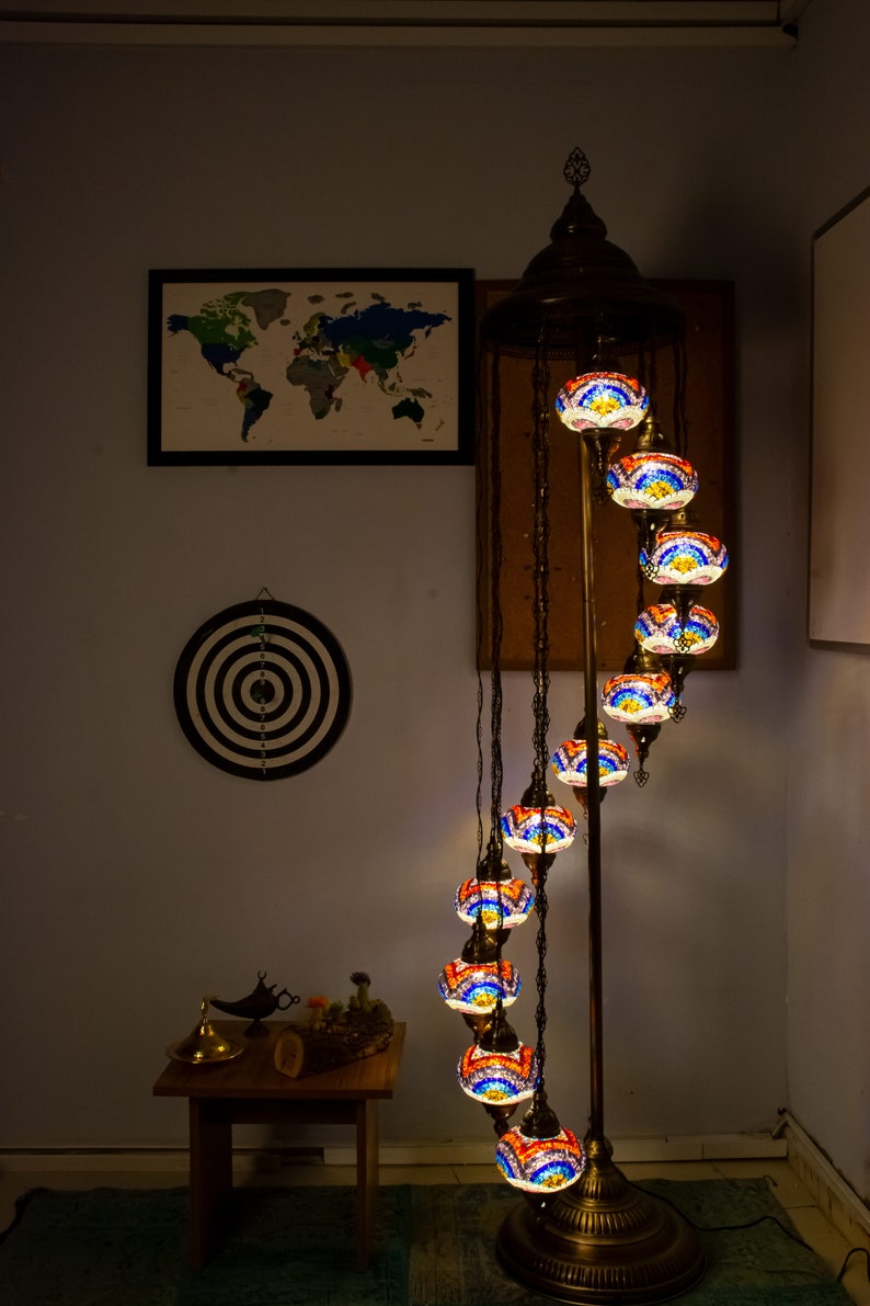 May include: A spiral floor lamp with a gold metal base and nine colorful glass globes. The globes are decorated with mosaic tiles in shades of blue, red, and orange. The lamp is lit and casting a warm glow.