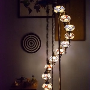 May include: A floor lamp with a curved design features a series of round, colorful glass globes. The globes are illuminated with warm white light. The lamp has a gold metal base and is standing on a dark brown rug.