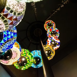May include: A collection of colorful, mosaic-style glass lanterns hanging from a metal pole. The lanterns are round and feature intricate patterns in shades of blue, green, red, and white.