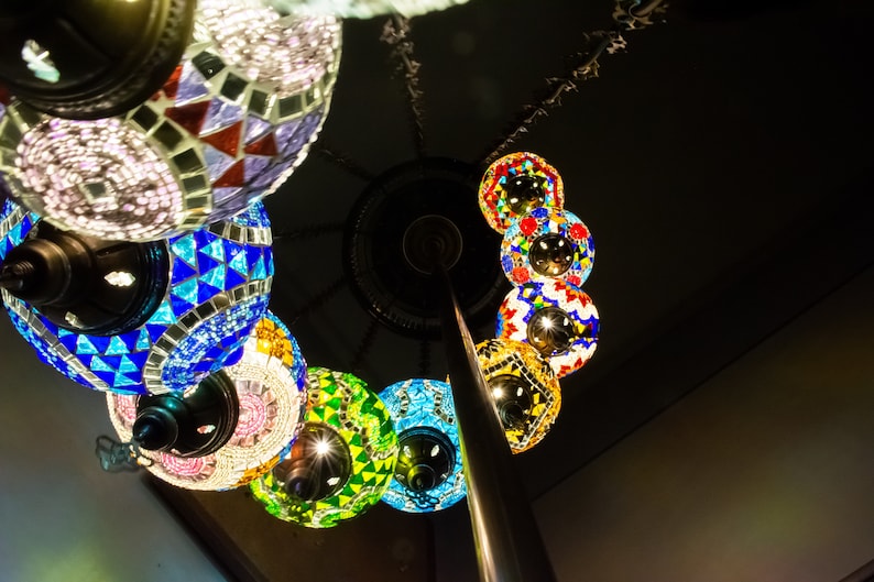May include: A collection of colorful, mosaic-style glass lanterns hanging from a metal pole. The lanterns are round and feature intricate patterns in shades of blue, green, red, and white.
