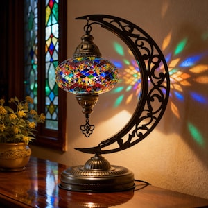May include: A decorative table lamp with a colorful mosaic glass shade. The lamp features a crescent-shaped metal frame with intricate cut-out designs. The lamp's base and accents have a bronze finish, and the light casts colorful patterns.