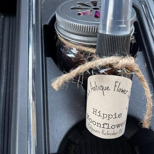 May include: A small glass bottle with a brown liquid and a white label that reads "Antique Flower" and "Hippie Moonflower Potpourri Refresher Oil". The bottle is tied with twine and is sitting in a car's cup holder next to a glass jar with a metal lid.