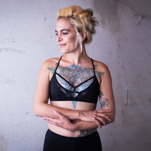 May include: A woman with blonde hair and tattoos is wearing a black sports bra with a mesh design and black pants. She has her arms crossed in front of her.