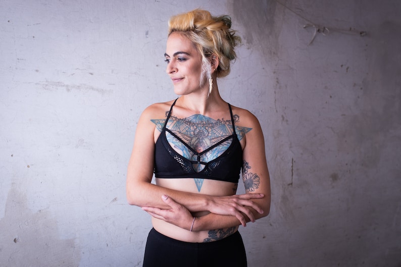 May include: A woman with blonde hair and tattoos is wearing a black sports bra with a mesh design and black pants. She has her arms crossed in front of her.