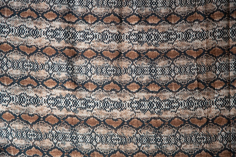 May include: A fabric with a snakeskin pattern. The design features a repeating pattern of brown, black, and cream-colored diamond shapes, creating a textured, reptile-like appearance. The fabric is likely used for clothing or accessories.