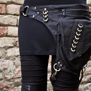 Puede incluir: Un bolso de cinturón utilitario negro con múltiples compartimentos con cremallera y un bolsillo grande con solapa. El bolso está adornado con anillos y remaches de metal plateado, con correas ajustables y clips que aseguran la bolsa al muslo.