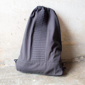 Puede incluir: Una mochila de cordón gris oscuro que presenta un panel vertical de pliegues horizontales en el centro. La bolsa tiene un cierre superior fruncido con cordones a juego y una sección inferior reforzada, confeccionada en tela sólida.
