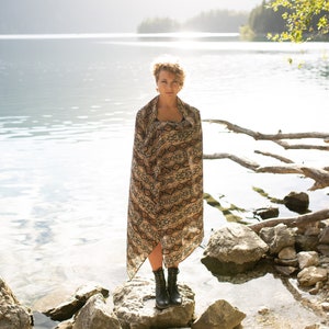 May include: A woman stands on rocks at the edge of a lake, wrapped in a patterned shawl. The shawl features a brown and tan snakeskin design. She wears black boots. The background includes the lake, trees, and sunlight.