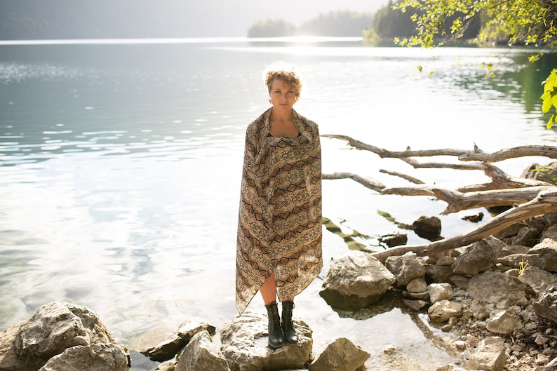 May include: A woman stands on rocks at the edge of a lake, wrapped in a patterned shawl. The shawl features a brown and tan snakeskin design. She wears black boots. The background includes the lake, trees, and sunlight.