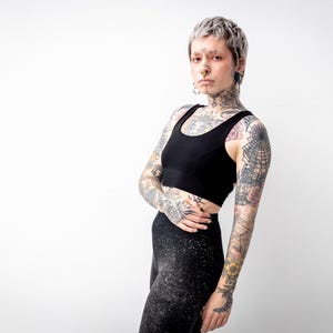 Könnte beinhalten: Ein schwarzer Sport-BH mit Rundhalsausschnitt und breitem Unterbrustband, kombiniert mit einer schwarzen Leggings mit dezentem, weißem Sprenkelmuster. Das Set wird von einer Person mit kurzem, silbernem Haar und vielen Arm- und Halstattoos getragen.