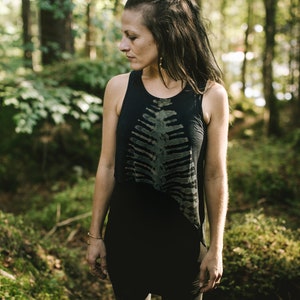Könnte beinhalten: Ein schwarzes ärmelloses Tanktop mit einem mittigen, dunkelgrünen Grafikdruck, der an einen Brustkorb erinnert. Das Kleidungsstück hat einen Rundhalsausschnitt und einen leicht asymmetrischen Saum, wobei sich das Motiv von der Brust bis zur Taille erstreckt.