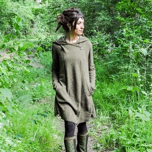 May include: A woman wearing a long, green hooded sweater with a zipper closure. She is standing in a forest setting.