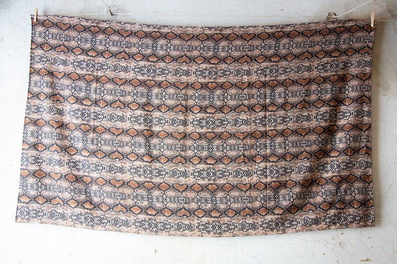 May include: A rectangular fabric with a snakeskin pattern. The design features a repeating pattern of brown, black, and tan shapes, resembling reptile scales. The fabric is hanging from a clothesline.
