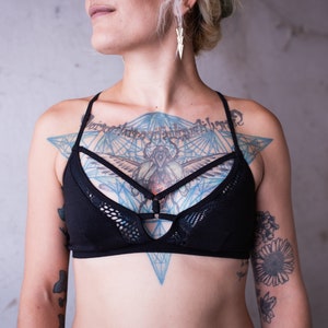 May include: A black lingerie top with a mesh overlay and a geometric design. The top features a criss-cross strap design and is worn over a chest tattoo.