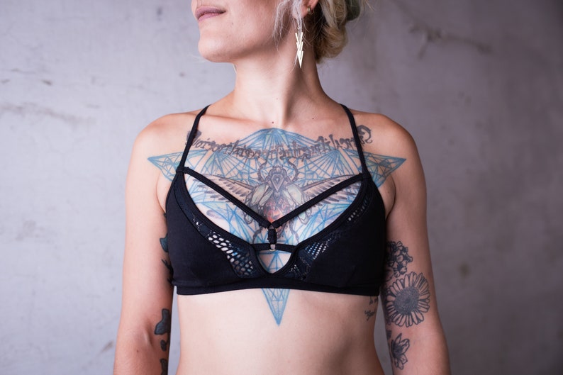 May include: A black lingerie top with a mesh overlay and a geometric design. The top features a criss-cross strap design and is worn over a chest tattoo.