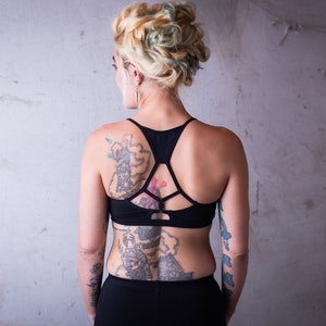 May include: A woman with blonde hair styled in a braid, wearing a black sports bra with a criss-cross back design. She has tattoos on her back, shoulders, and arms.