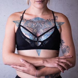 May include: A woman with a black bra with a mesh overlay and a geometric tattoo on her chest. The tattoo is blue and black and features a winged insect.