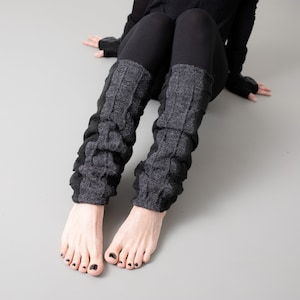 May include: A pair of grey and black knitted leg warmers with a cable knit pattern. The leg warmers are worn on a person's legs, with the feet bare.