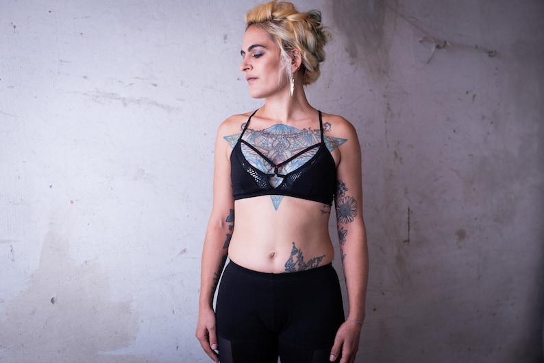 May include: A woman with short blonde hair and tattoos is wearing a black lace bralette with a geometric design. She is also wearing black leggings.