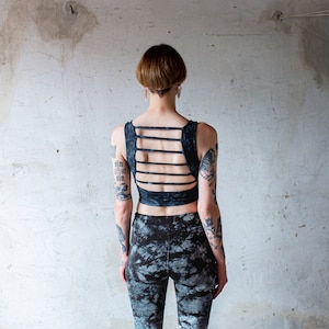 Puede incluir: Una persona con el pelo corto y oscuro y tatuajes lleva un top corto negro con un dise&ntilde;o de espalda cruzada y pantalones estampados en blanco y negro.