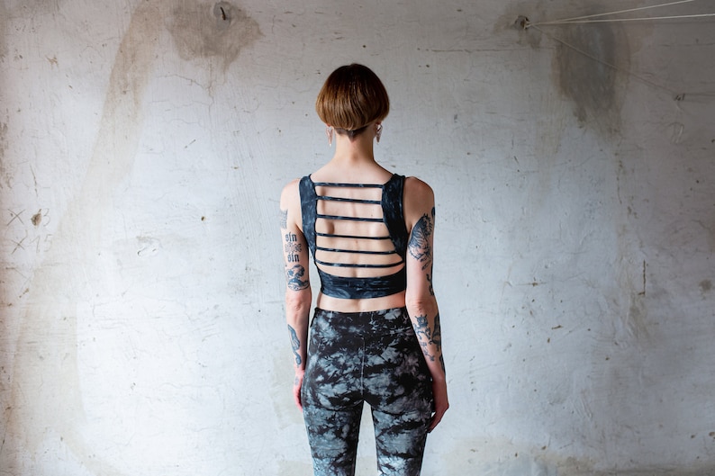 Puede incluir: Una persona con el pelo corto y oscuro y tatuajes lleva un top corto negro con un dise&ntilde;o de espalda cruzada y pantalones estampados en blanco y negro.