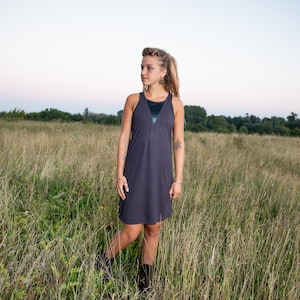 May include: A woman with long, braided hair stands in a field of tall grass wearing a dark gray sleeveless dress. She is looking off to the side.