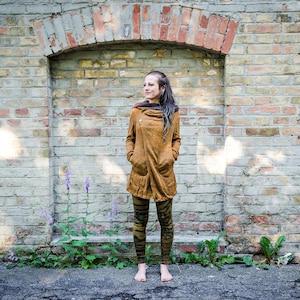 Könnte beinhalten: Eine Frau trägt eine braune Wildlederjacke mit Kapuze, schwarze und grüne gemusterte Leggings und barfuß. Sie steht vor einer Backsteinmauer mit einer Tür rechts.