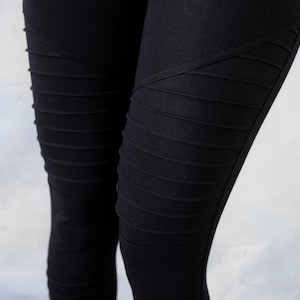 Könnte beinhalten: Schwarze Leggings mit einem markanten, vom Motorradstil inspirierten Design mit horizontalen Rippeneinsätzen an Oberschenkeln und Knien. Der Stoff ist einfarbig schwarz mit matter Oberfläche und zeigt eine eng anliegende Passform.