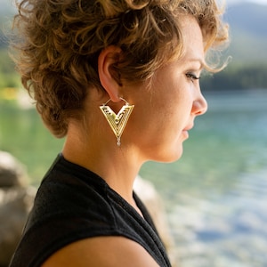 Könnte beinhalten: Goldfarbene geometrische Ohrringe in V-Form mit aufwendigen, geschichteten Designs. Die Ohrringe sind an einem Reifen befestigt und hängen leicht. Das Bild zeigt die Ohrringe am Ohr einer Person, mit einem verschwommenen Hintergrund aus See und Bäumen.