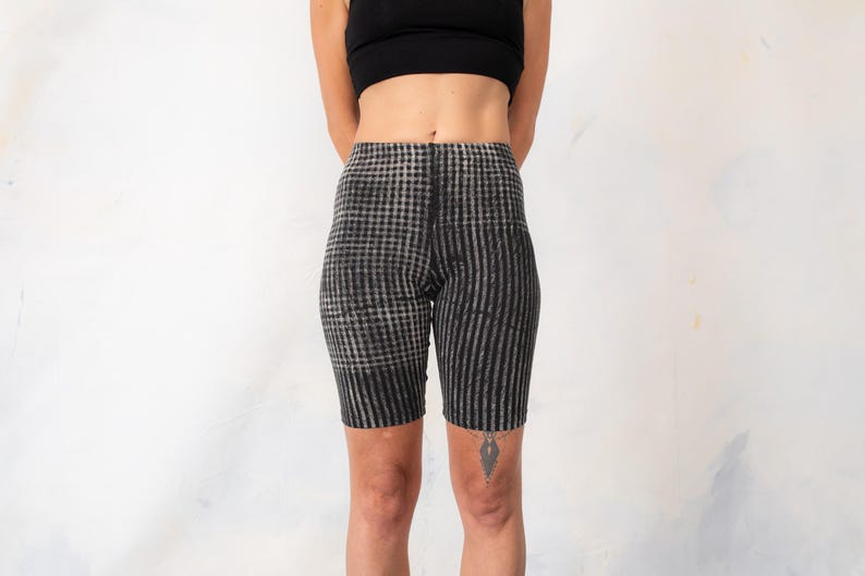 May include: High-waisted biker shorts featuring a black and grey pattern. The left leg displays a checkered design, while the right leg features vertical stripes. These form-fitting shorts reach mid-thigh and have a seamless, elasticized waistband.
