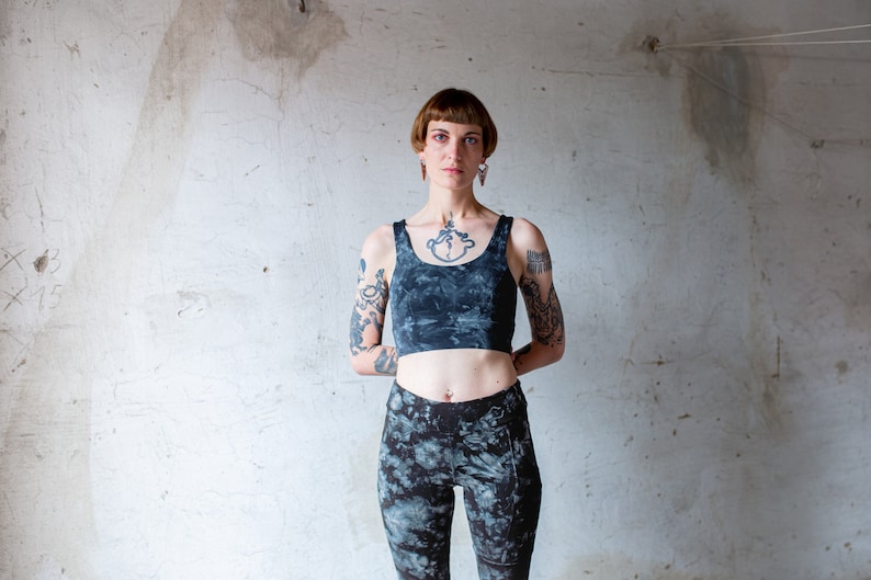 Puede incluir: Una mujer con el pelo corto y casta&ntilde;o oscuro y tatuajes en los brazos lleva un top corto azul oscuro y leggings a juego. El top tiene un estampado tie-dye y los leggings tienen un estampado similar.