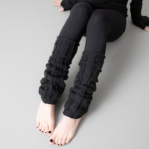 May include: Black knitted leg warmers with a textured pattern. The leg warmers are worn on a person's legs, with the feet bare.