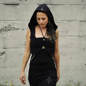 May include: A black hooded vest with a black dress underneath. The vest has gold accents and a zipper on each side. The model is wearing the vest and dress with the hood up.