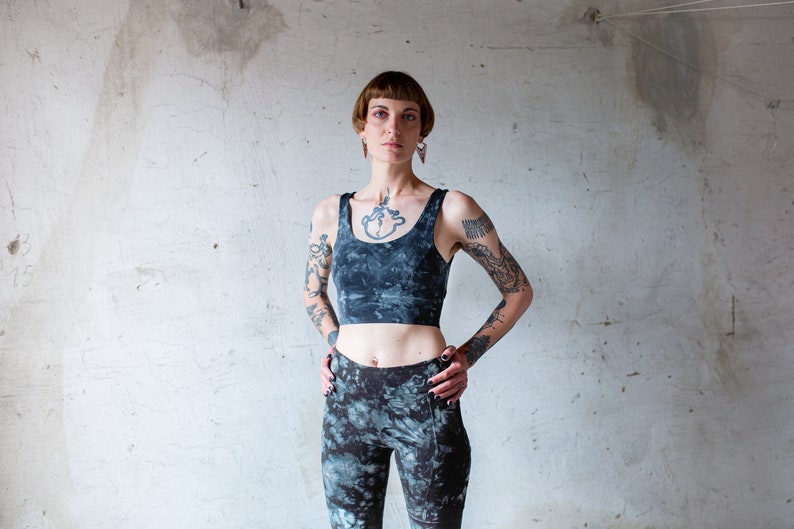 Puede incluir: Una mujer con el pelo corto y oscuro y tatuajes lleva un sujetador deportivo de color negro y gris tie-dye y leggings a juego. Est&aacute; de pie frente a una pared blanca.