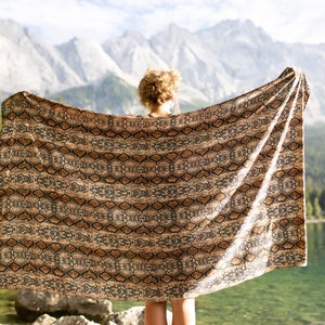 May include: A large, rectangular blanket with a snakeskin pattern in shades of brown and tan is held up against a backdrop of mountains, a lake, and trees. The blanket is draped, showcasing its design and texture.